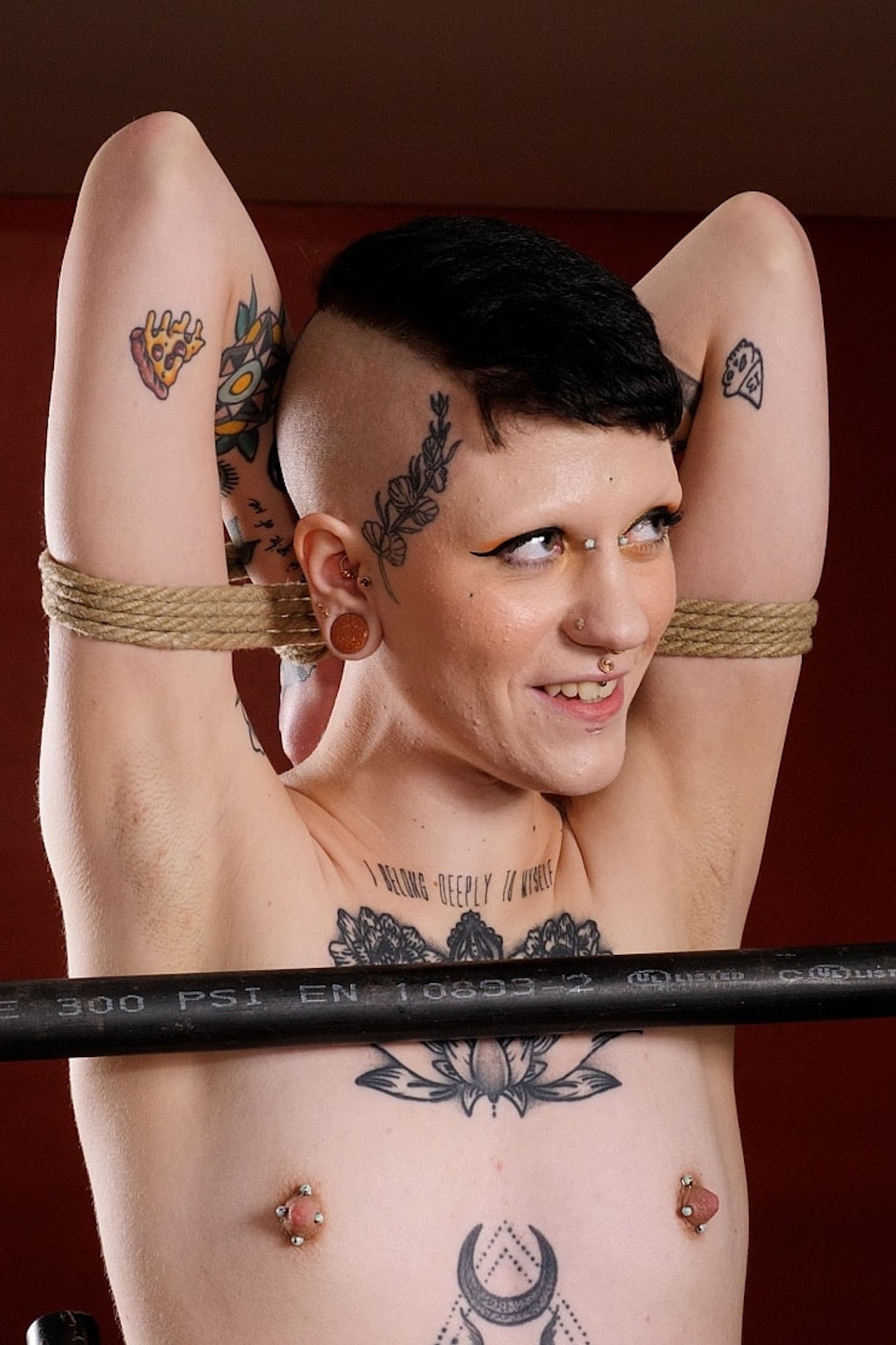 Portrait of naked tattooed person with small breasts and nipple piercings. They are tied up.